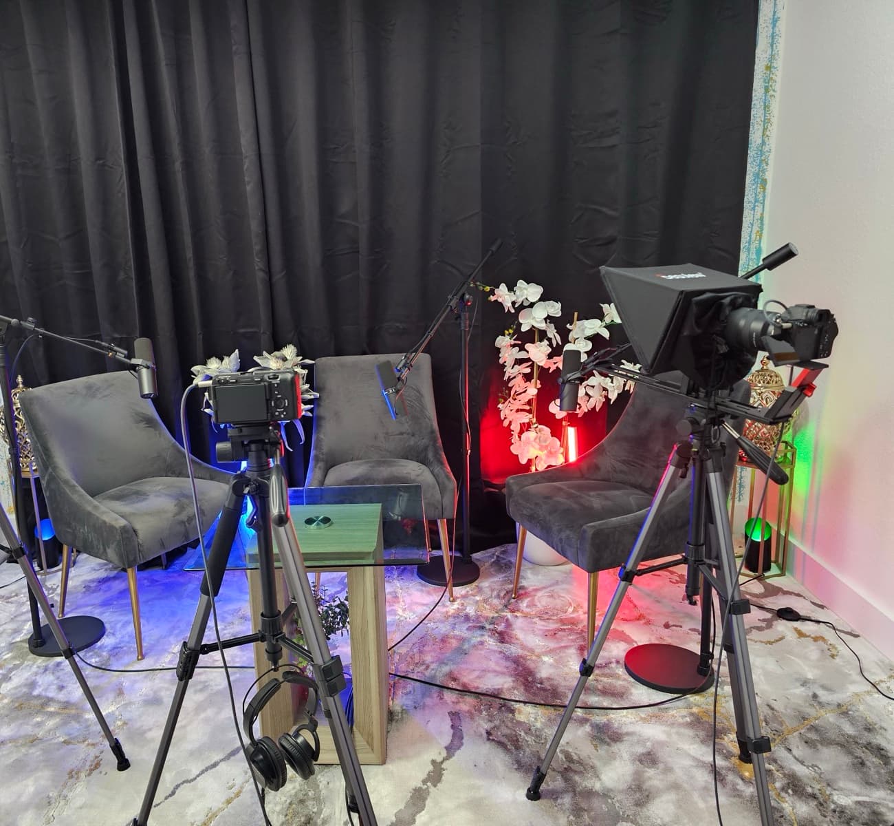 Two tripod-mounted cameras, one fitted with a teleprompter, facing the three-chair podcast set against a black curtain backdrop