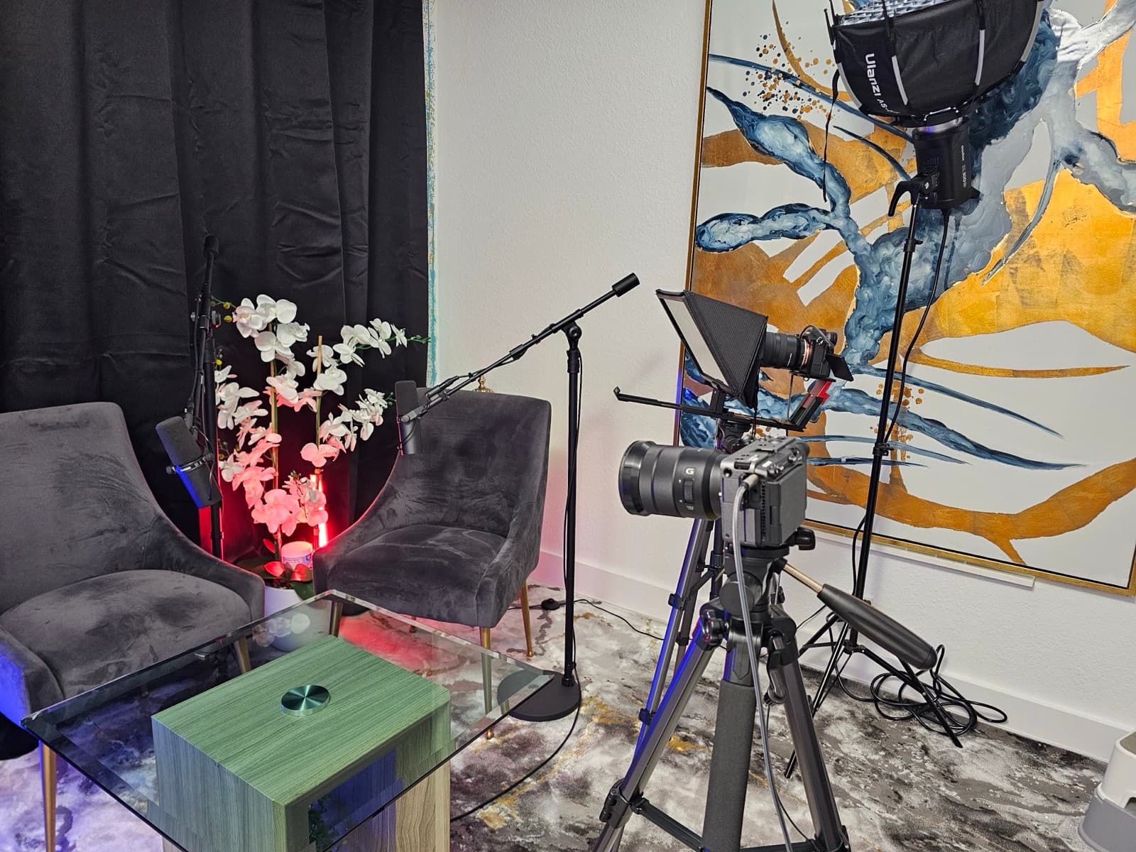 Teleprompter-equipped camera and softbox studio light set up beside a large blue and gold abstract painting, with velvet chairs and orchids nearby