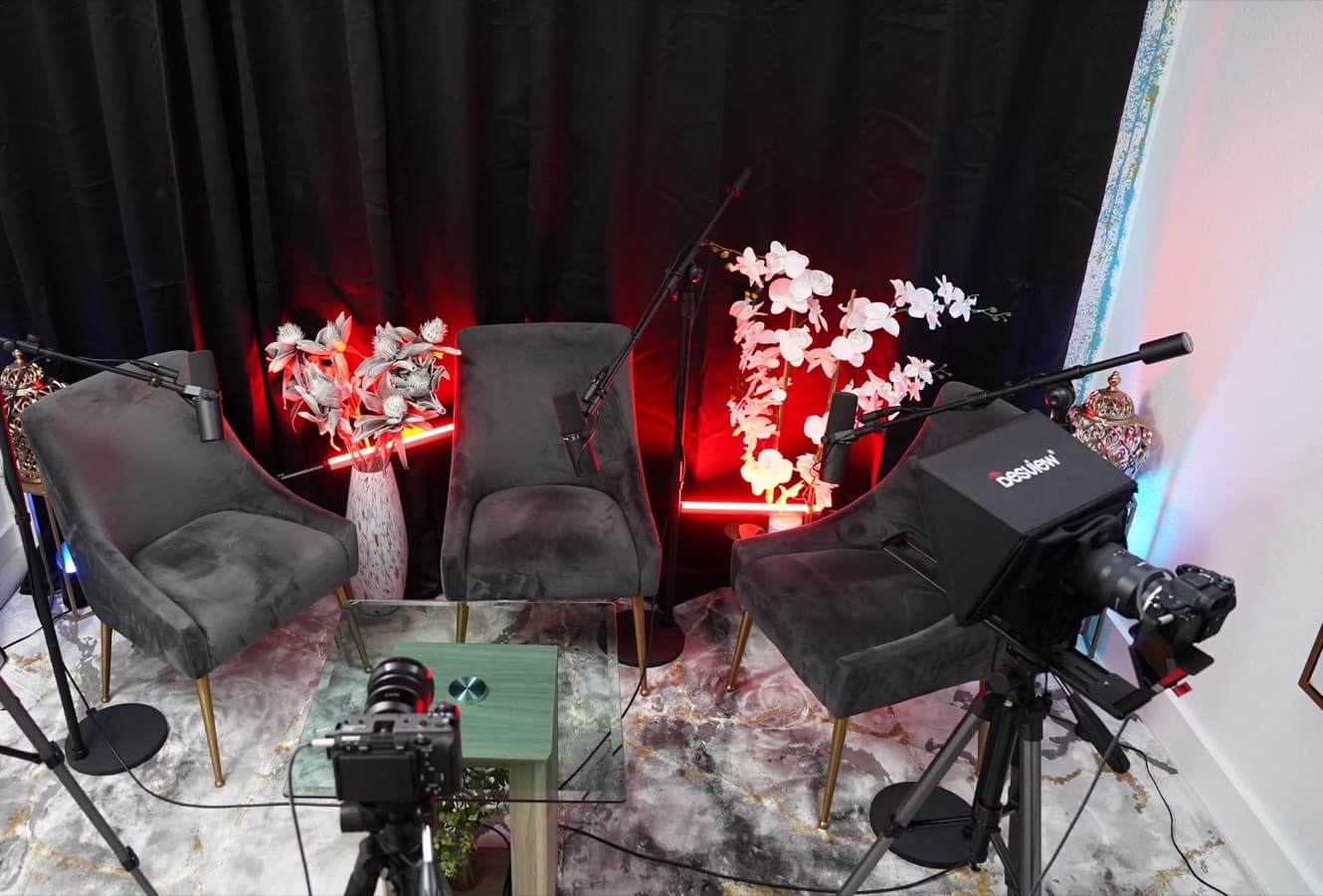 Elevated view over the camera rigs onto the three-chair podcast set with glass table, orchids, and red LED accent lighting