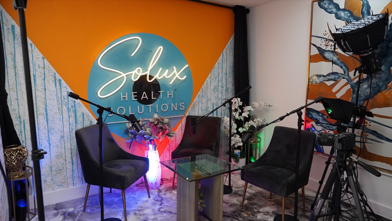 Wide view of the Solux Studios set showing the neon sign, three chairs, boom microphones, teleprompter camera, and softbox studio light beside large wall art