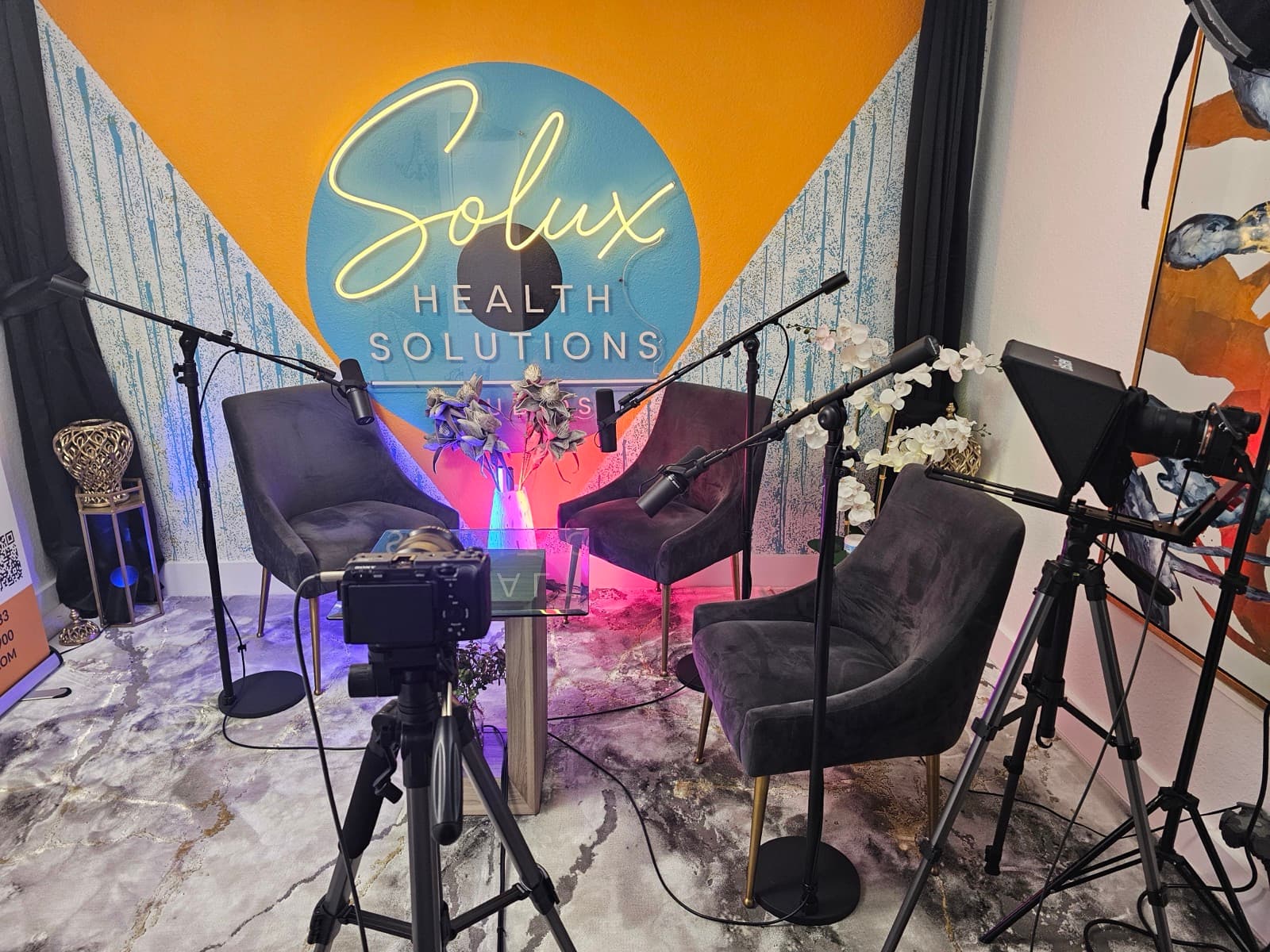 Straight-on view of the Solux Studios recording set with the neon sign centered above three velvet chairs and a glowing accent vase
