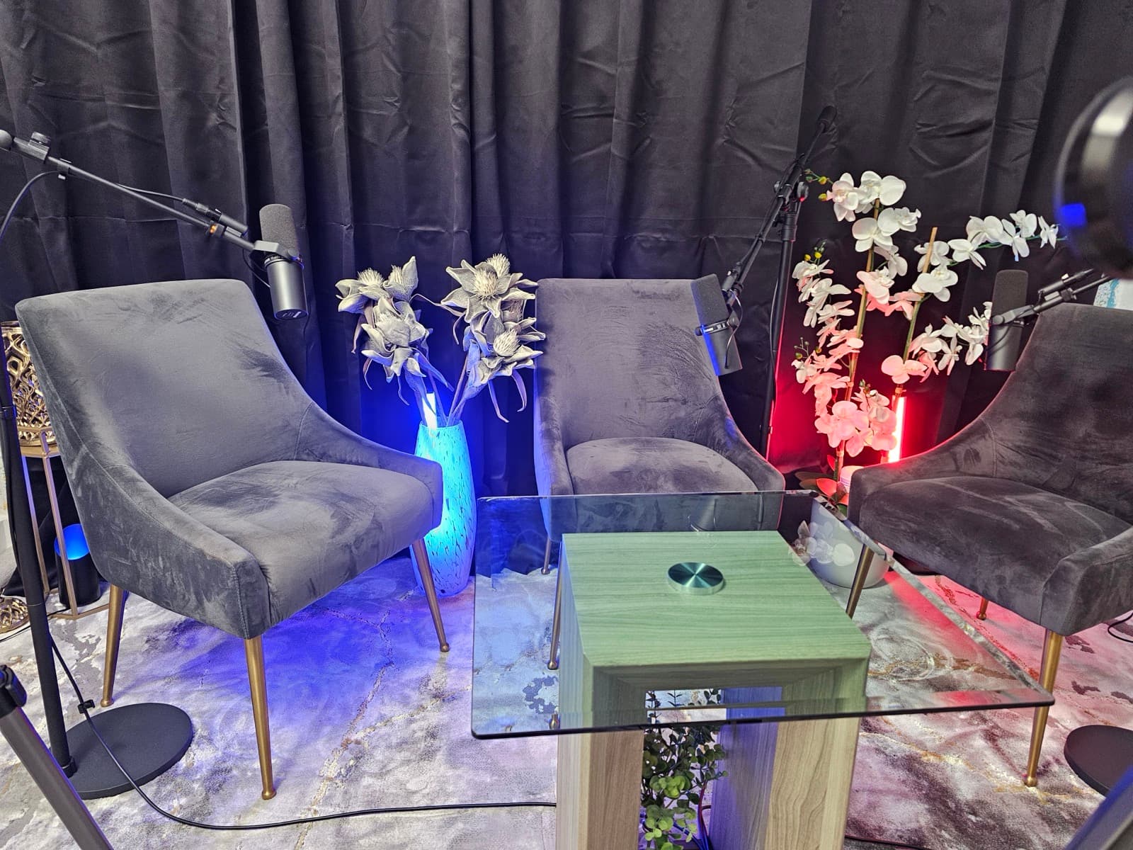 Straight-on view of three velvet chairs with boom-mounted microphones around a glass coffee table, with orchids and a dark curtain backdrop