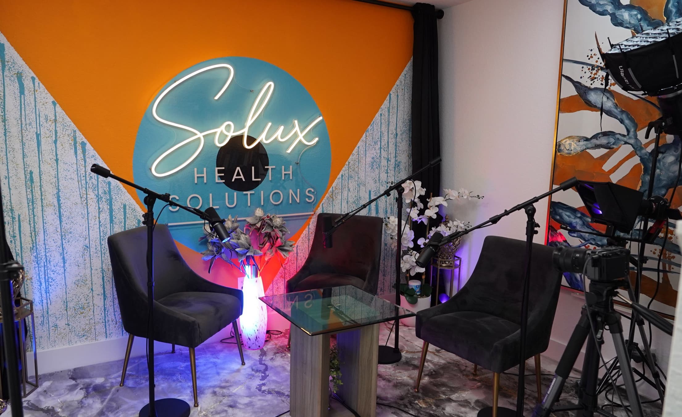 Wide view of the Solux Studios podcast set with a glowing neon sign on an orange and teal mural wall, three grey velvet chairs around a glass table, and studio microphones